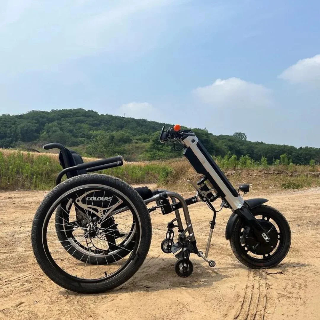 The product in the image is an electric attachment for a wheelchair, model Mijo MT01. The image shows the electric attachment connected to a wheelchair, allowing for easier mobility. The attachment has two large wheels and one smaller caster, providing stability and maneuverability. In the background is a natural landscape with greenery and a blue sky.