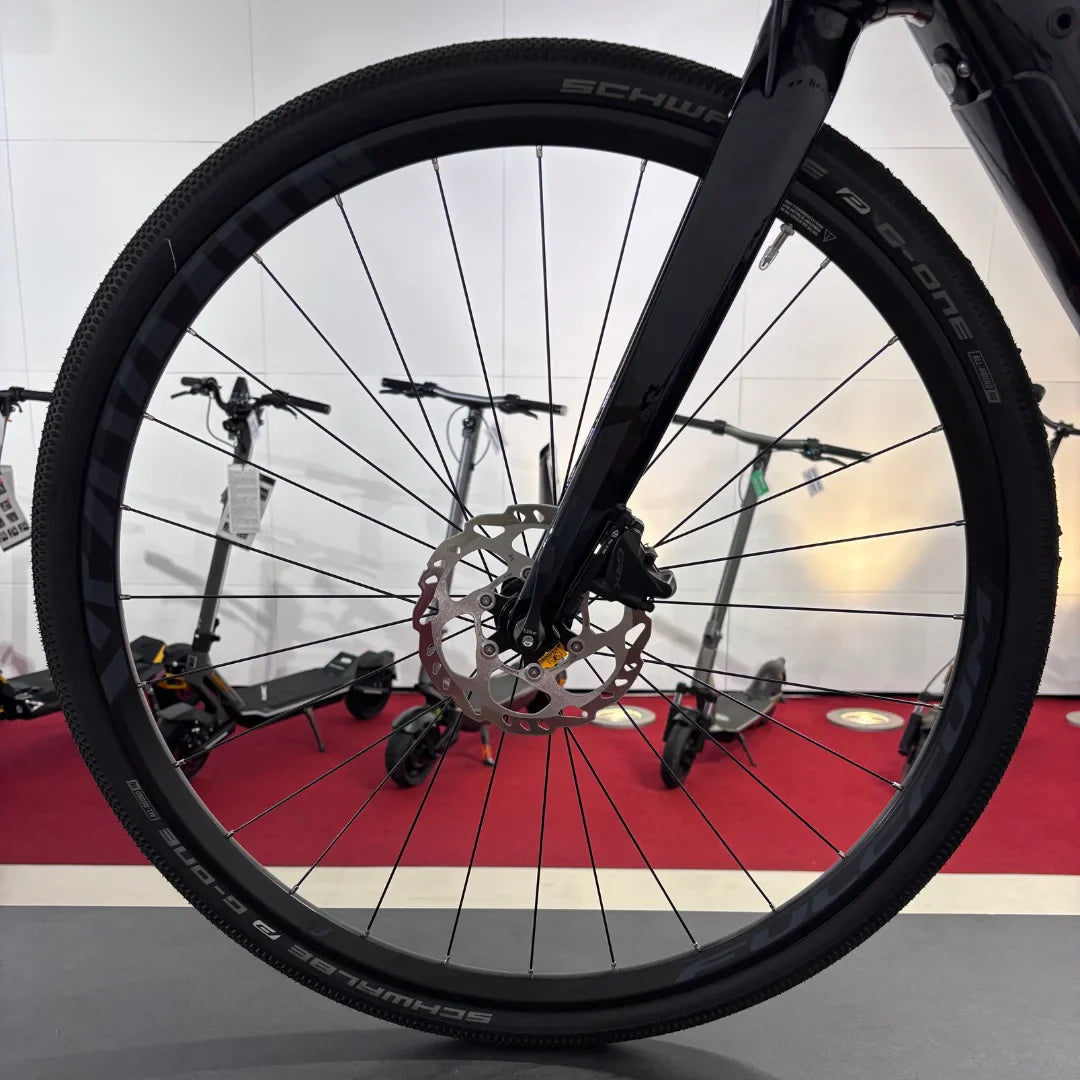 The product in the image is an Electric Bike, brand Raymon, model GravelRay E 7.0. The image shows the front wheel with black tires and disc brakes, indicating a sporty design. Electric scooters are also visible in the background, giving a sense of the variety of transport options available.