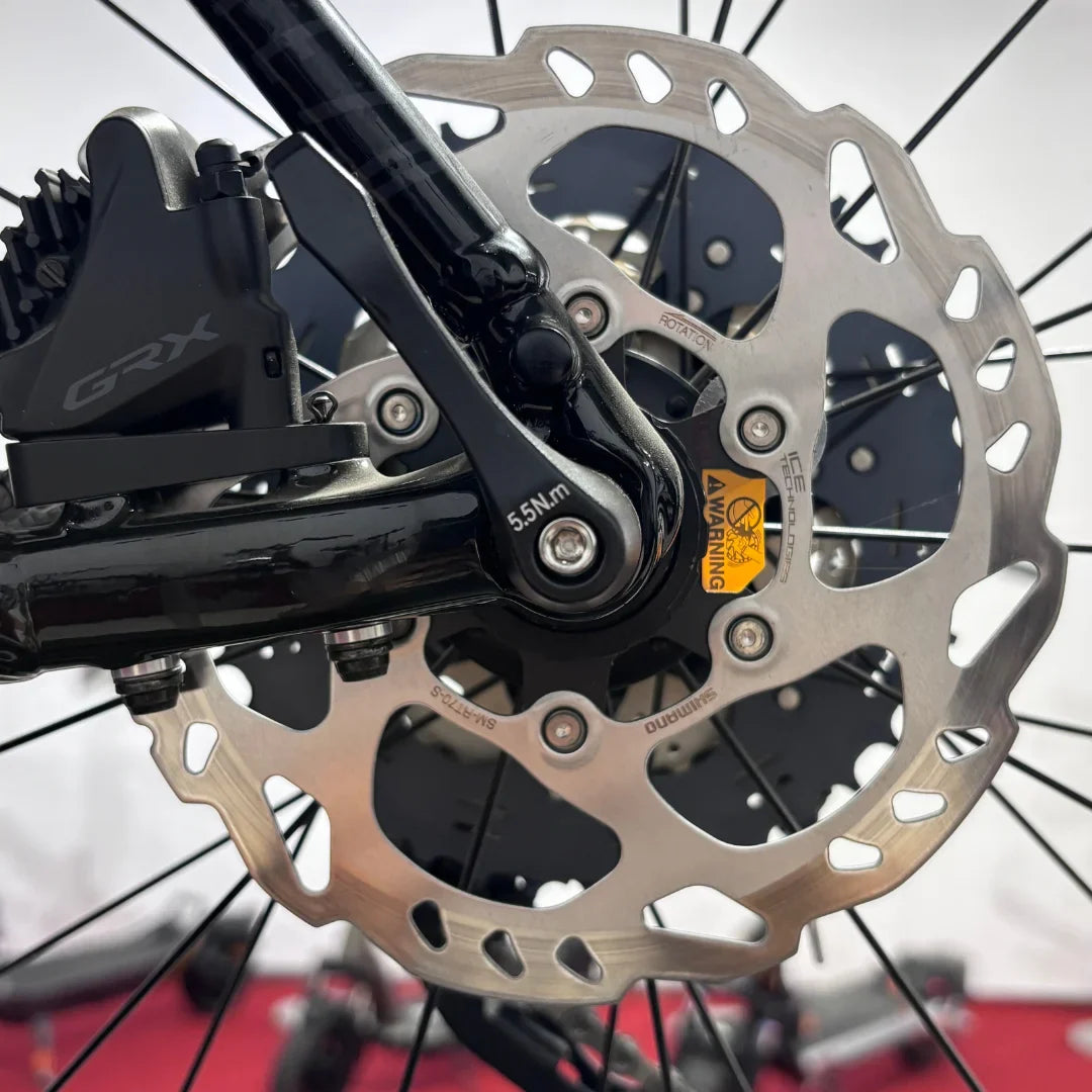 The product is an Electric Bike, brand is Raymon, and the model is GravelRay E 7.0. The image shows a part of the rear wheel, where the brake disc and shifting system are visible. The brake disc is made of stainless steel, ensuring good braking performance, while a marking for correct installation is visible on the frame.