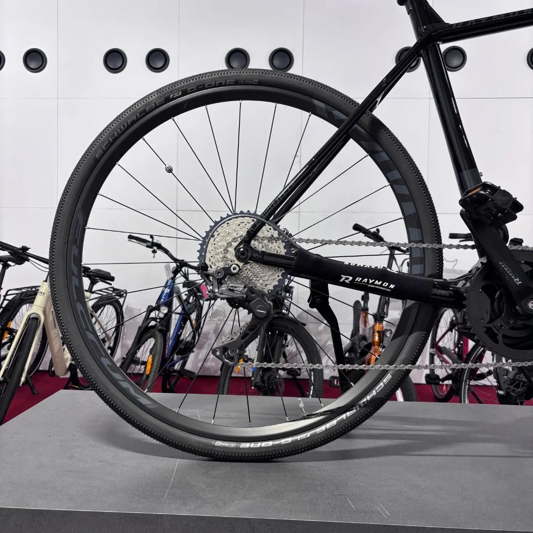 The product in the image is an electric bike, model Raymon GravelRay E 7.0. The image shows the rear wheel with tires suitable for gravel terrain, and part of the drivetrain, including the cassette and chain. The bike is on a stand, with other bikes visible in the background.