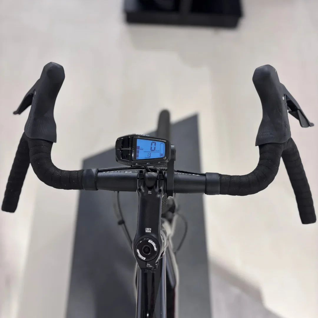 The product pictured is an Electric Bike, model Raymon GravelRay E 7.0. The image shows handlebars with rubber grips and a digital display showing speed and other data. The bike is designed for gravel riding, meaning it is suitable for various terrains.