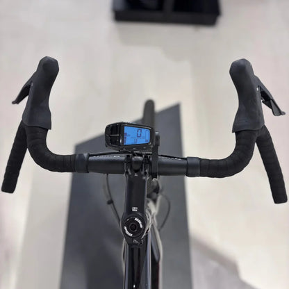 The product pictured is an Electric Bike, model Raymon GravelRay E 7.0. The image shows handlebars with rubber grips and a digital display showing speed and other data. The bike is designed for gravel riding, meaning it is suitable for various terrains.
