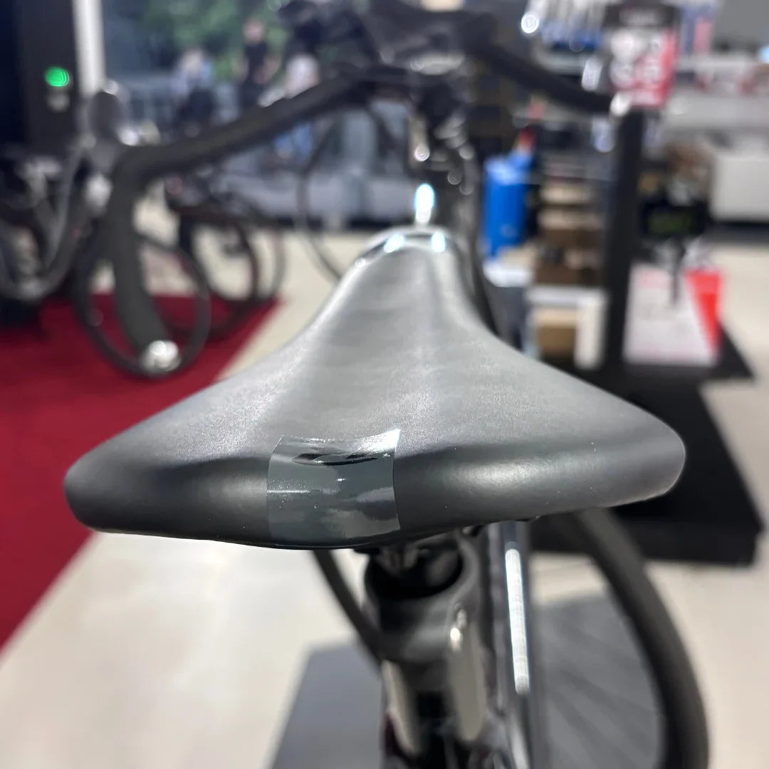 The product pictured is an electric bike, brand Raymon, model GravelRay E 7.0. The image shows the bike's seat, which is black and has adhesive tape on it, indicating possible damage or repair. In the background, part of the bike and the store's surroundings are visible.