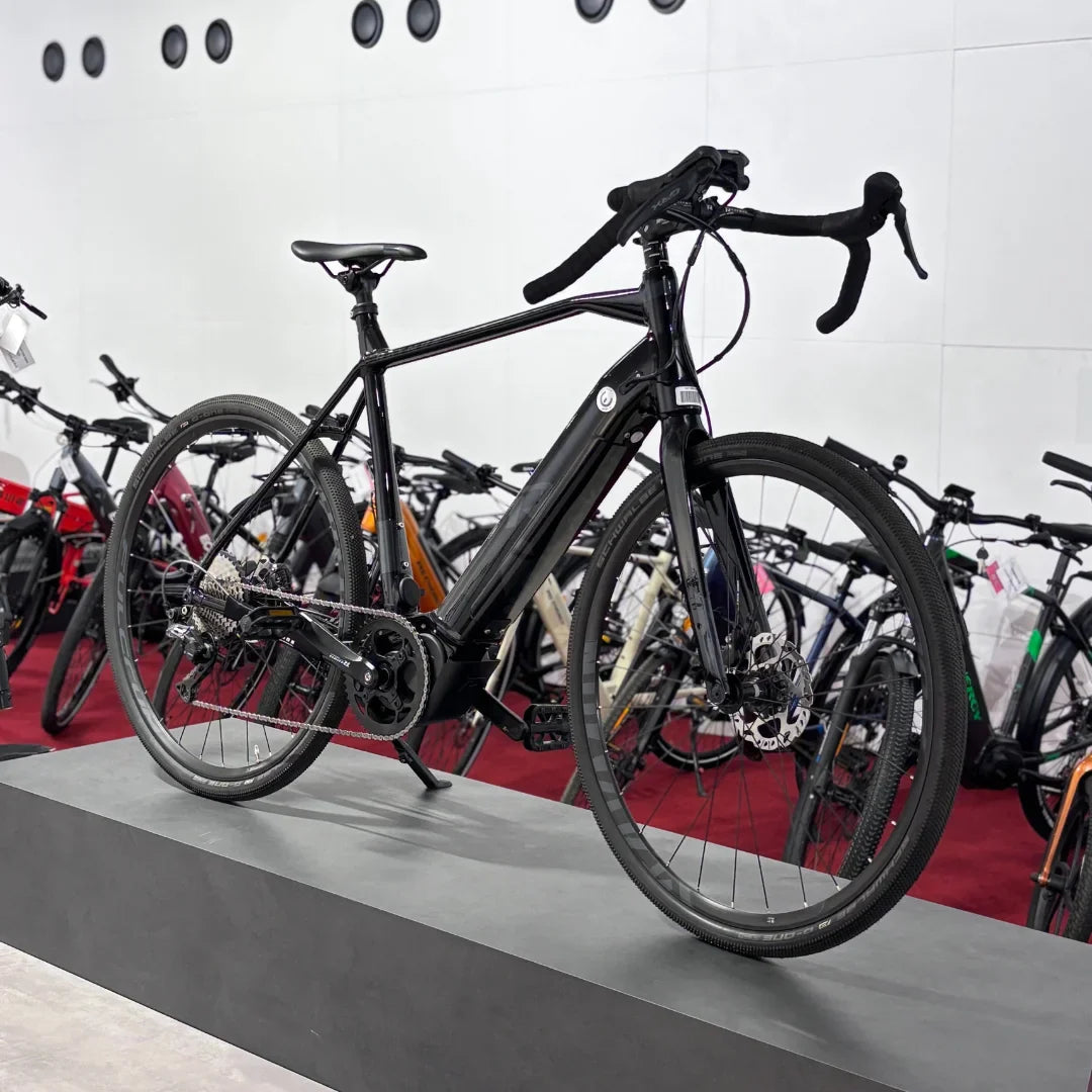 The product pictured is an electric bike, brand Raymon, model GravelRay E 7.0. The bike is black and has a sporty design, typical of gravel bikes. In the picture, it is placed on a stand, with other bikes visible in the background. The bike features wide tires and disc brakes, which provide good grip and braking.