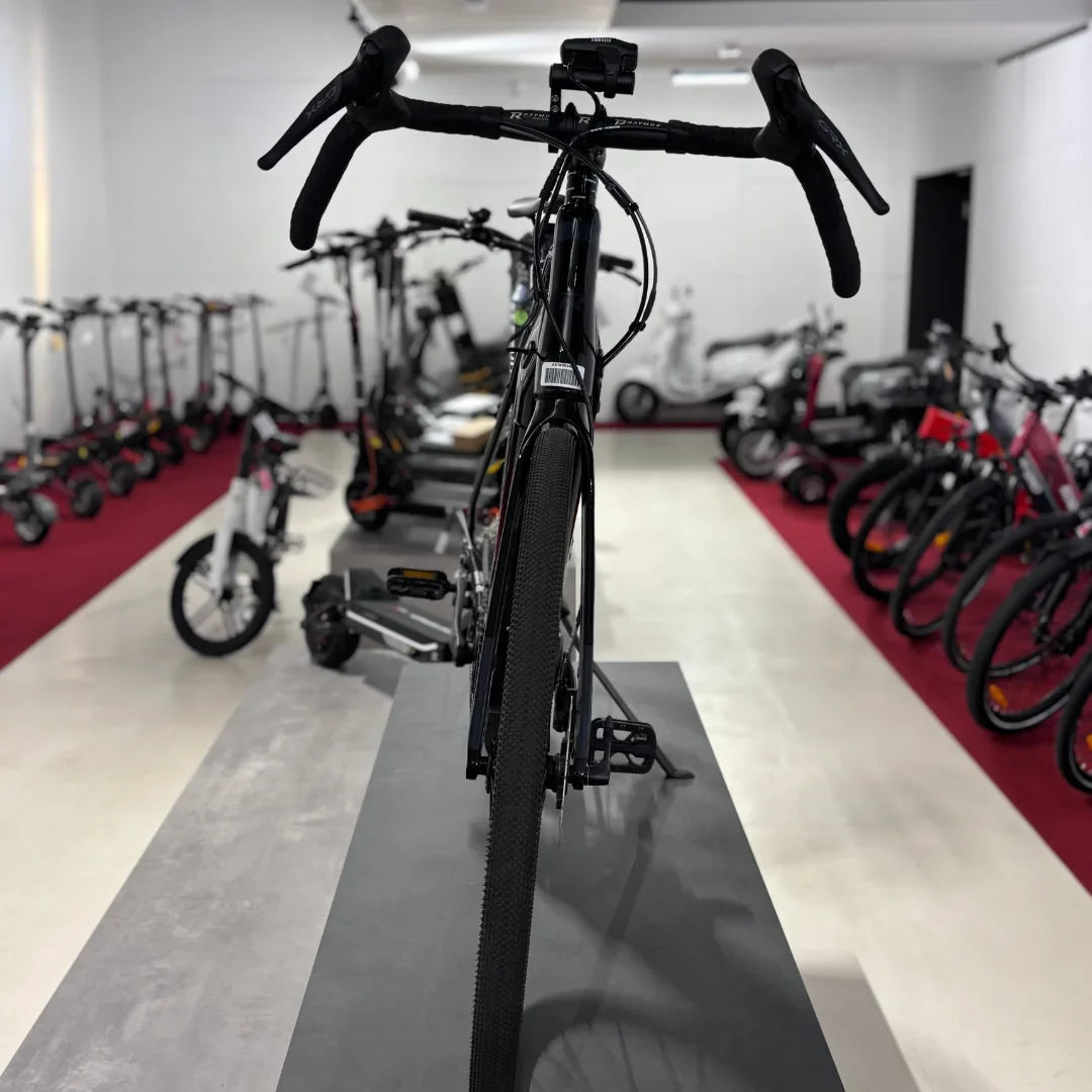 The product in the image is an electric bike, specifically the Raymon GravelRay E 7.0 model. The image shows the back of the bike with visible handlebars and wide tires, indicating its suitability for gravel terrains. Other electric scooters and bikes are also visible in the background, suggesting a retail environment.