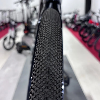 The product pictured is an electric bike, brand Raymon, model GravelRay E 7.0. The image shows the bike's profile with a special tire texture designed for riding on various terrains. Other cycling equipment is visible in the background, suggesting the picture was taken in a store or exhibition.