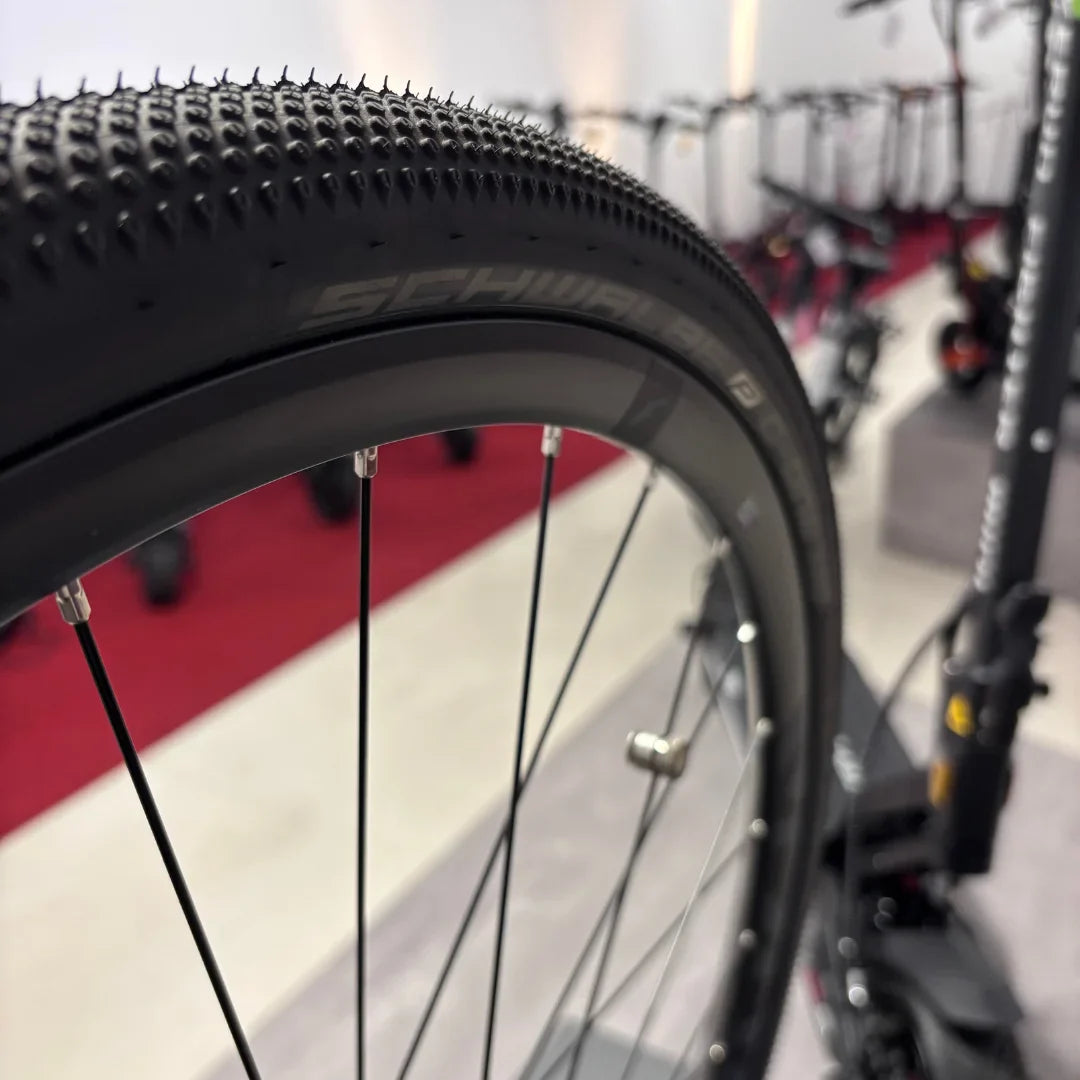 The product is an Electric Bike, brand Raymon, model GravelRay E 7.0. The image shows a part of the bike, specifically a tire with a tread suitable for gravel surfaces, along with a part of the rim and spokes. Other bikes are also visible in the background, suggesting an exhibition or store setting.