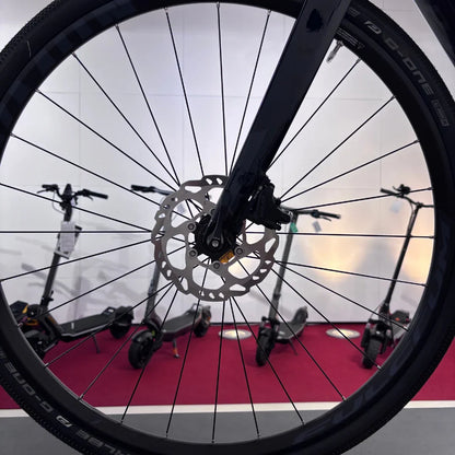 The product in the image is an electric bike, brand Raymon, model GravelRay E 7.0. The front part of the bike is shown, with the rim, spokes, and disc brakes visible, while electric scooters are visible in the background. The bike is designed for riding on various terrains.