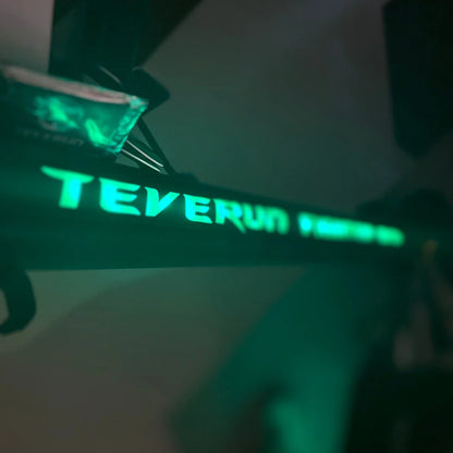 Izdelek na sliki je električni skiro, model Teverun Fighter Mini Pro iz leta 2025. Na sliki je vidna osvetljena oznaka znamke Teverun na okvirju skiroja, ki daje sodoben in atraktiven videz. V ozadju je tudi del skiroja, kar nakazuje na njegovo zasnovo in funkcionalnost.