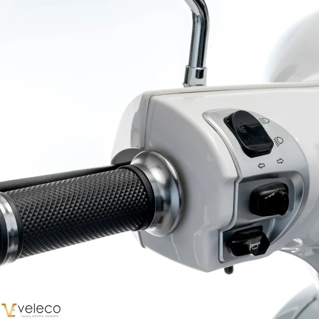 The image shows an Electric Motorcycle, model Veleco Cristal. The handlebars and part of the control panel are visible, with buttons for lights and turn signals. The motorcycle has an elegant design with a smooth white surface and textured rubber grips for a comfortable hold.