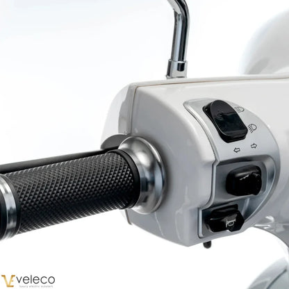 The image shows an Electric Motorcycle, model Veleco Cristal. The handlebars and part of the control panel are visible, with buttons for lights and turn signals. The motorcycle has an elegant design with a smooth white surface and textured rubber grips for a comfortable hold.