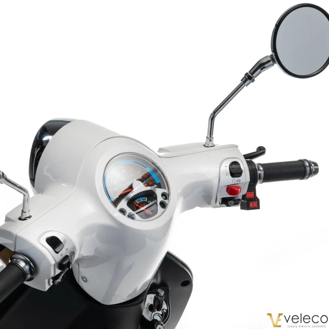 The product is an Electric motorcycle, brand Veleco, model Cristal. The image shows the control panel of the motorcycle, which includes the speedometer, control levers, and mirror. The color combination is white and black, giving the motorcycle an elegant look. Control buttons and details for easy use are also visible.