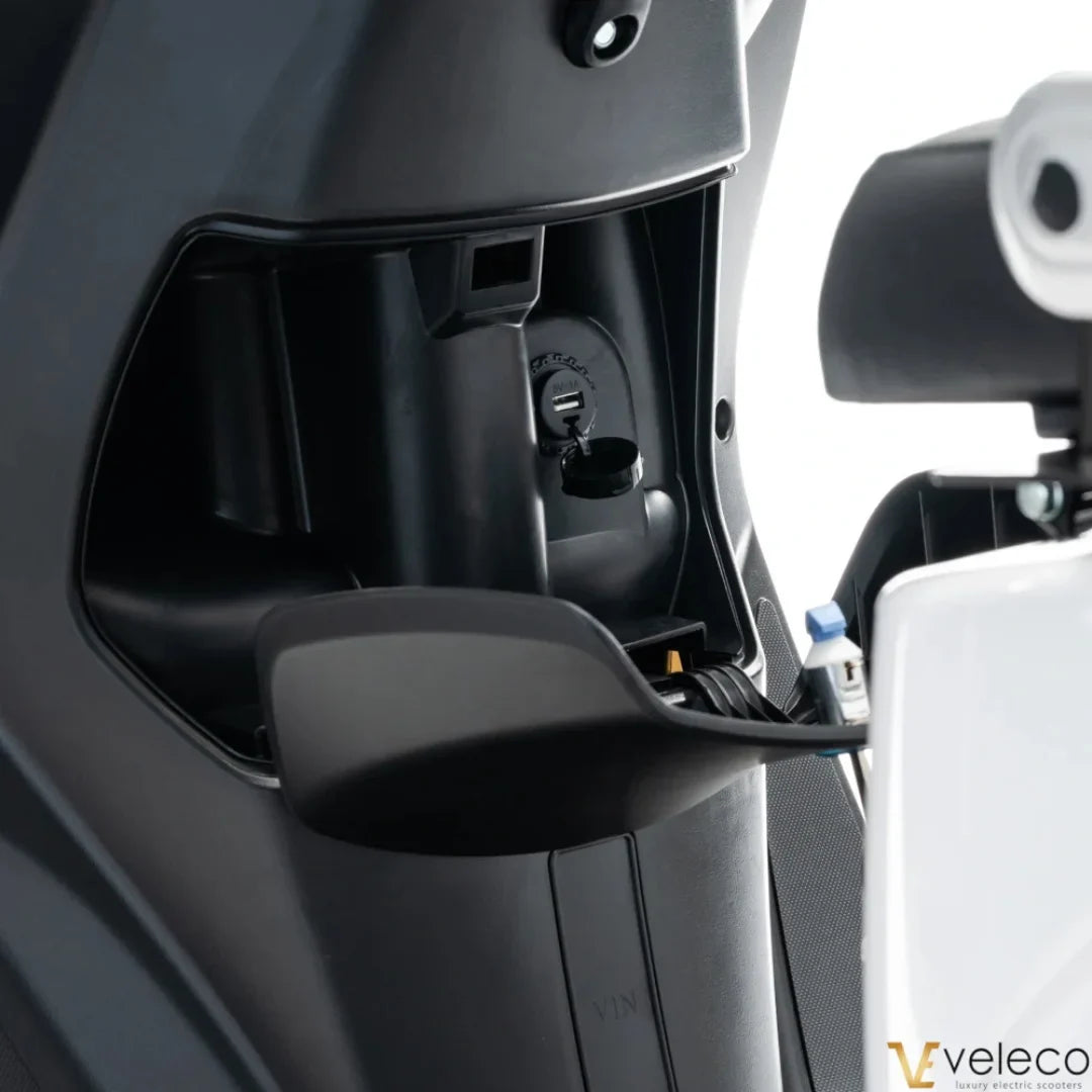 The product in the image is an Electric Motorcycle from the brand Veleco, model Cristal. The image shows part of the scooter's interior, revealing connectors and operational mechanisms, indicating a modern design and functionality. The equipment also includes a charging port, which is crucial for the electric vehicle's operation.