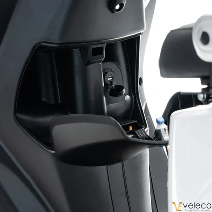 The product in the image is an Electric Motorcycle from the brand Veleco, model Cristal. The image shows part of the scooter's interior, revealing connectors and operational mechanisms, indicating a modern design and functionality. The equipment also includes a charging port, which is crucial for the electric vehicle's operation.