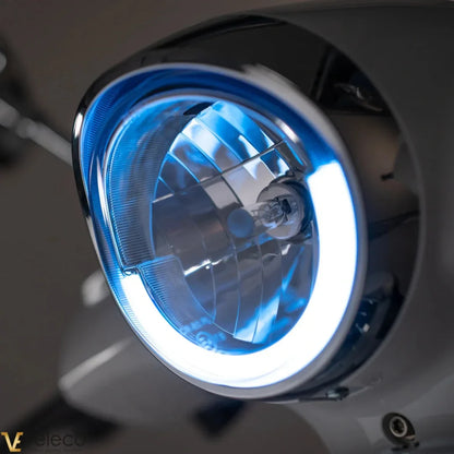 The product in the image is an Electric motorcycle, brand Veleco, model Cristal. The image shows the scooter's front light, which features blue illumination and a modern design. The light is designed to enhance visibility and safety while riding.