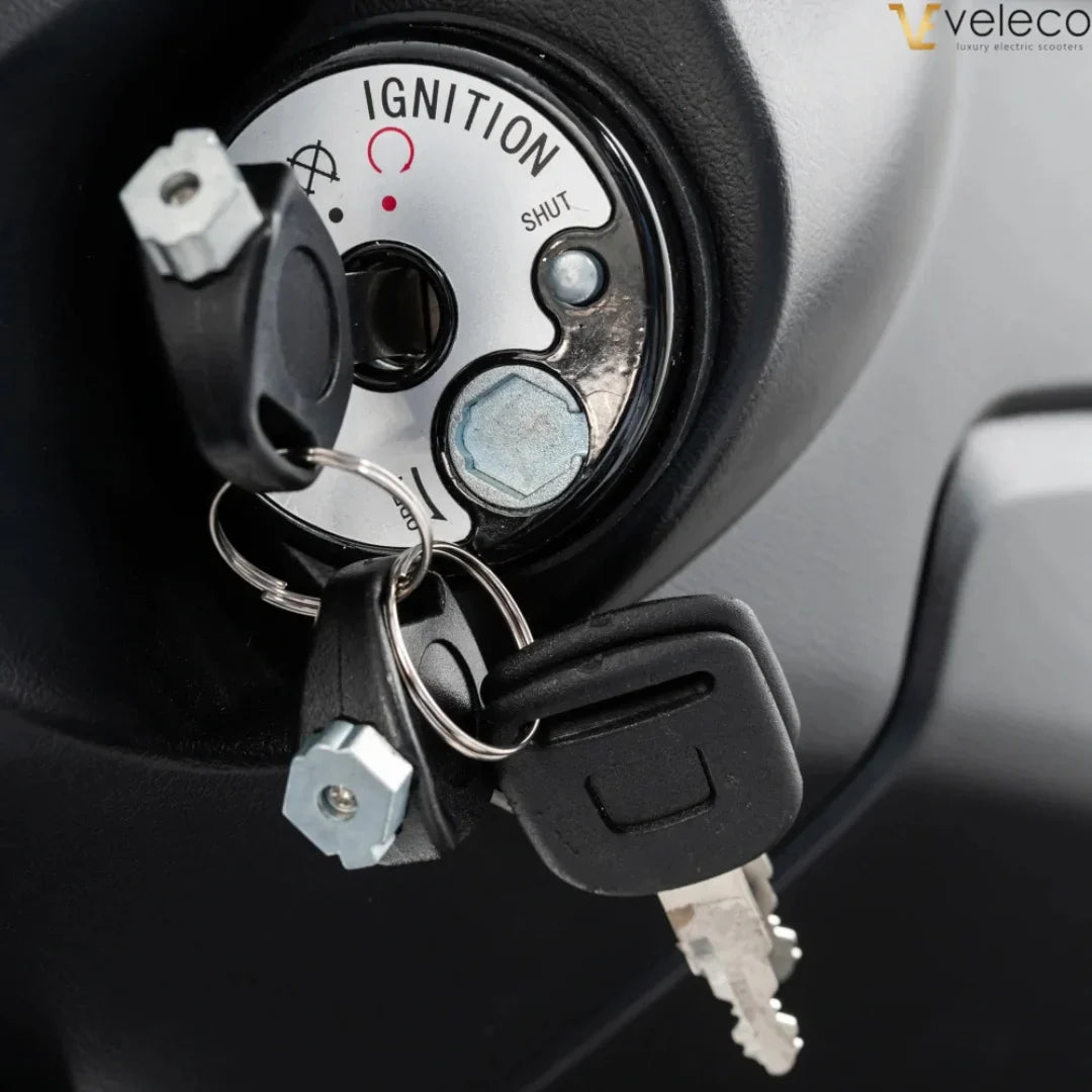 The product is an Electric motorcycle, brand Veleco, model Cristal. The image shows the ignition lock, which includes keys and operating markings such as "SHUT" and "IGNITION". The construction around the lock is also visible, indicating the scooter's functionality and design.