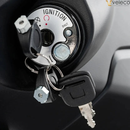 The product is an Electric motorcycle, brand Veleco, model Cristal. The image shows the ignition lock, which includes keys and operating markings such as "SHUT" and "IGNITION". The construction around the lock is also visible, indicating the scooter's functionality and design.
