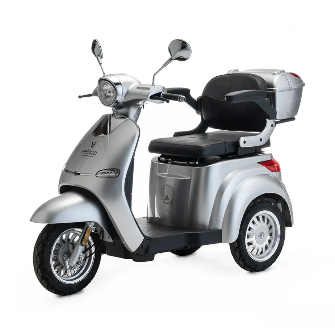 The product pictured is an Electric mobility scooter by Veleco, model Cristal. It is a three-wheeled electric scooter designed for comfortable and safe riding. The image shows its silver body, a comfortable seat, and a rear storage basket for easily stowing personal items.