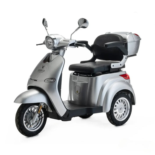 The product pictured is an Electric mobility scooter by Veleco, model Cristal. It is a three-wheeled electric scooter designed for comfortable and safe riding. The image shows its silver body, a comfortable seat, and a rear storage basket for easily stowing personal items.