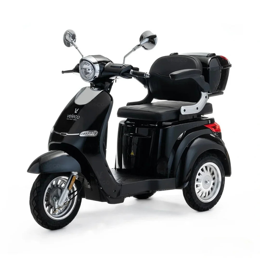 The product is an Electric motorcycle, model Veleco Cristal. The image shows a black, three-wheeled electric vehicle equipped with a comfortable seat and a trunk at the rear. The motorcycle features a classic design with retro elements, including chrome mirrors and a front light.