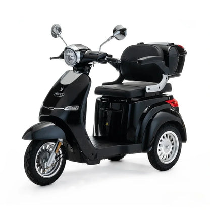The product is an Electric motorcycle, model Veleco Cristal. The image shows a black, three-wheeled electric vehicle equipped with a comfortable seat and a trunk at the rear. The motorcycle features a classic design with retro elements, including chrome mirrors and a front light.