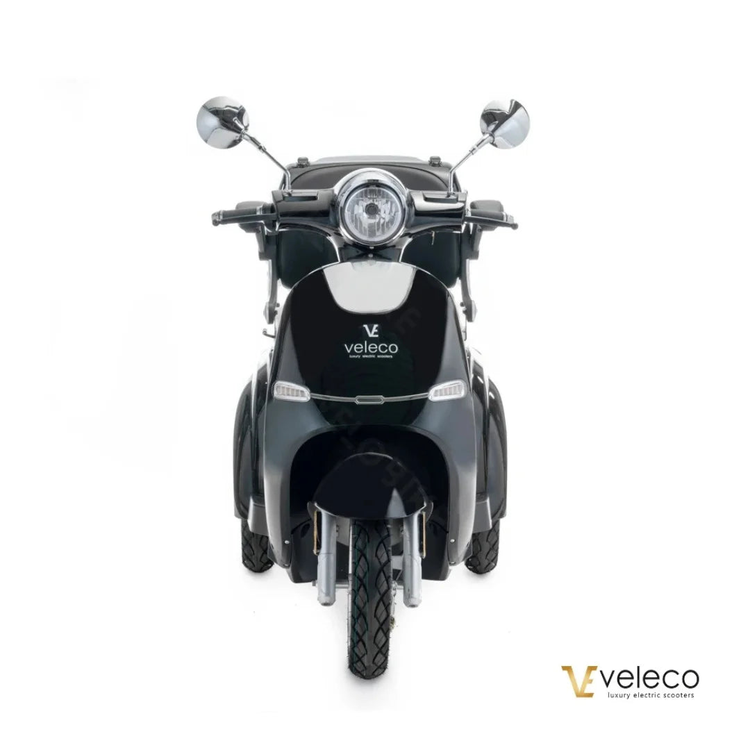 The product is an Electric motorcycle of the brand Veleco, model Cristal. The image shows the front of the scooter, which has a retro design with distinctive chrome mirrors and a round front light. The scooter's color is dark, giving it an elegant look. The Veleco brand name is clearly visible on the rear.