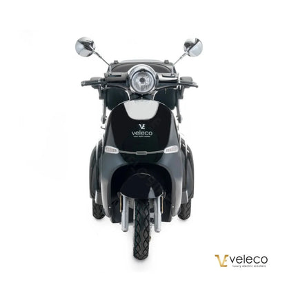 The product is an Electric motorcycle of the brand Veleco, model Cristal. The image shows the front of the scooter, which has a retro design with distinctive chrome mirrors and a round front light. The scooter's color is dark, giving it an elegant look. The Veleco brand name is clearly visible on the rear.