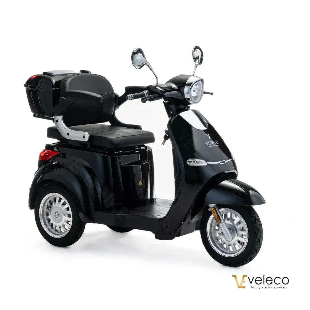The product pictured is an electric scooter, brand Veleco, model Cristal. The scooter features a sleek black design, a comfortable seating area, and is equipped with front and rear mirrors and a storage compartment at the back. It has three wheels, ensuring stability and easy handling.