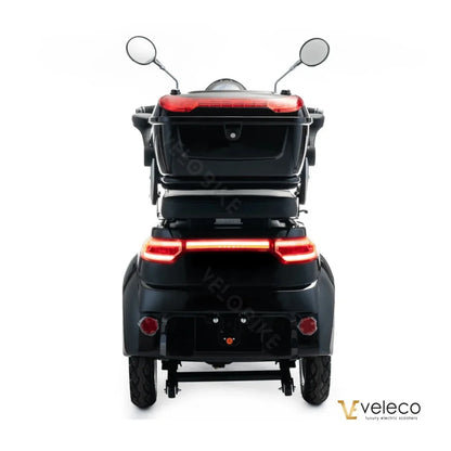The image shows an Electric motorcycle, model Veleco Cristal. The product has three wheels for stable riding and is equipped with mirrors and taillights. The motorcycle is black and includes storage space, enhancing its functionality.