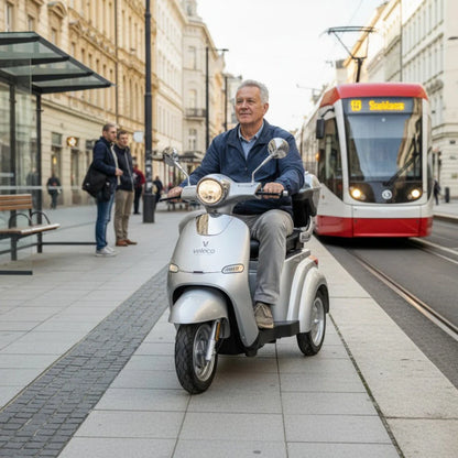 The product in the image is an electric scooter, model Veleco Cristal. The image shows a man riding the scooter in an urban environment, with a tram and pedestrians nearby. The scooter has a modern design and is equipped with lighting elements, conveying a sense of comfort and mobility in city traffic.