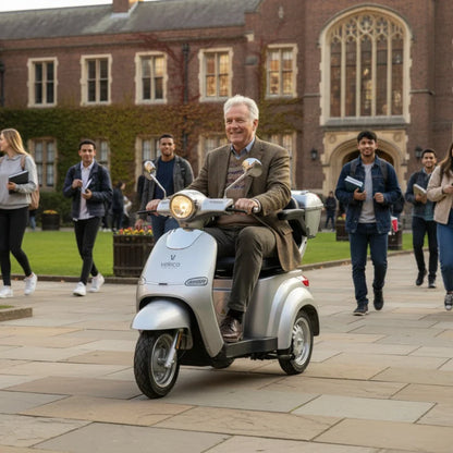 The image shows an Electric motorcycle from the brand Veleco, model Cristal. The product is silver and has a retro design. An elderly man is sitting on the scooter, smiling as he rides through a university courtyard, surrounded by young people walking around. Buildings with distinctive architecture are visible in the background, giving the image a pleasant and lively atmosphere.